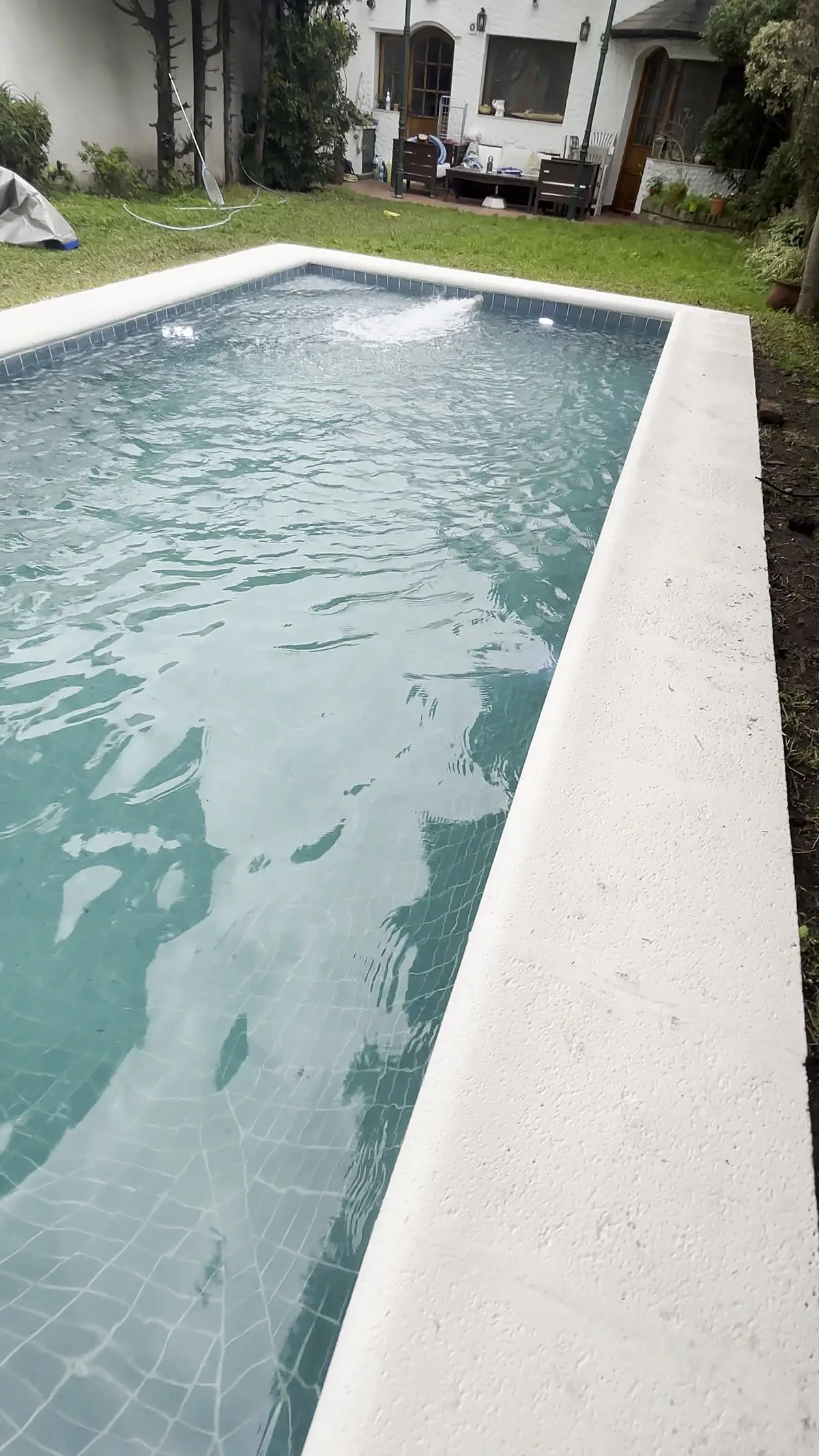 Piscina reformada y llena, con el borde blanco y el jardín alrededor